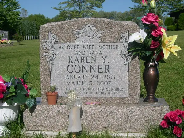 A light grey granite upright monument with a carved cherub above the name, lilies cut at both shoulders and a scripture line across the foot