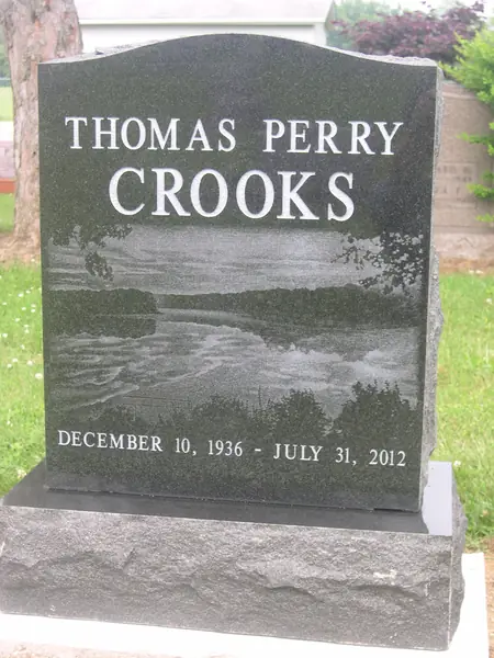 A polished black granite upright monument with a full-width etched river and woodland scene beneath the name, on a rock-pitched base