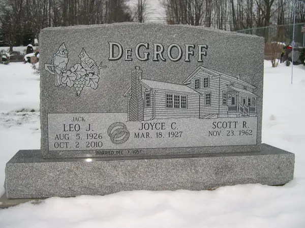 A grey granite upright monument with an etched house, cardinals and a grapevine, three name tablets and a wedding-ring emblem, standing in snow