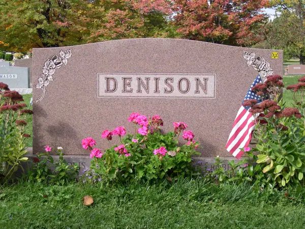 A rose granite upright monument with a raised name panel and carved flowers at both upper corners, with a veterans marker and flag at the base