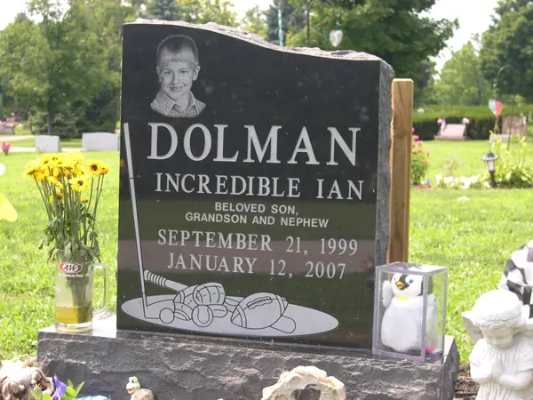 A polished black granite upright monument with a rough-hewn top edge, a laser-etched portrait of a boy and etched sports equipment below the name