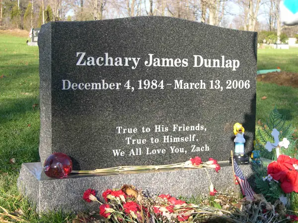 A polished dark grey granite upright monument with a shaped top, the full name and dates on one line and a three-line epitaph below