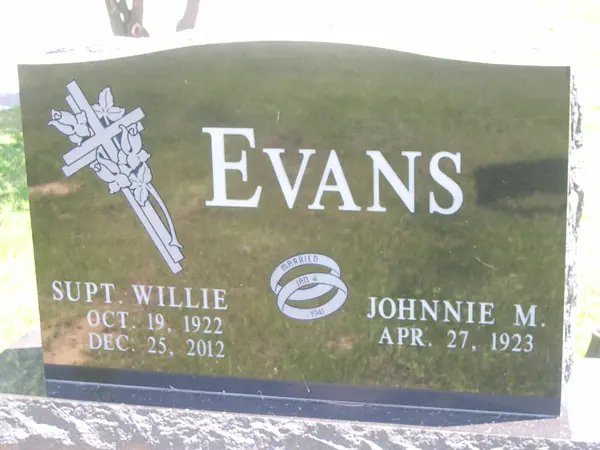 A polished dark granite upright monument with a serpentine top, a carved cross wrapped in lilies at one shoulder and a wedding-ring emblem between the name tablets