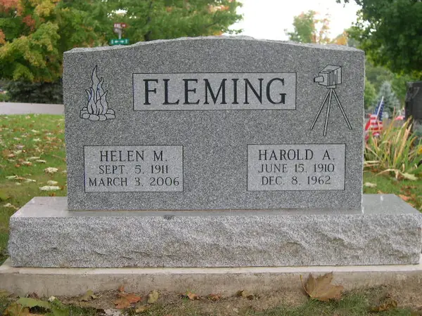 A grey granite upright monument with a raised name panel, a campfire carved at one shoulder and a camera on a tripod at the other