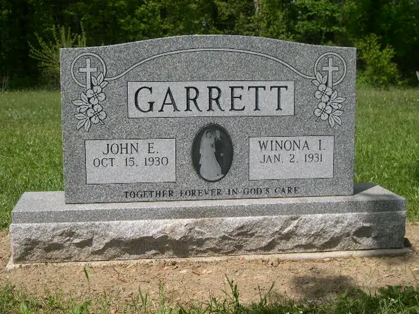 A grey granite upright monument with carved crosses and flowers at both shoulders, an etched oval wedding portrait between the name tablets and an epitaph across the foot