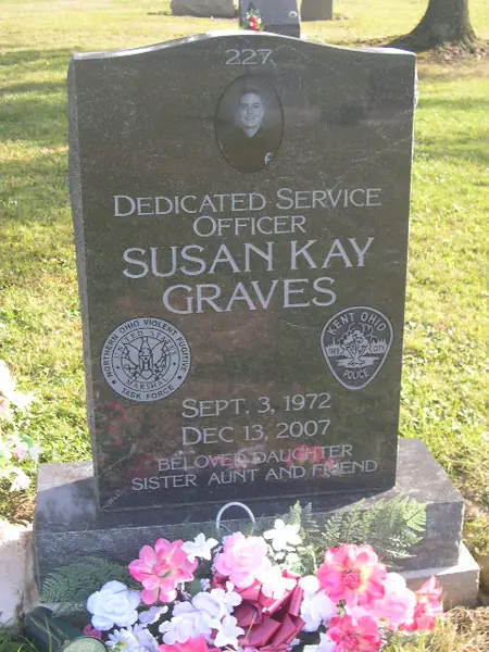 A dark granite upright monument with an etched oval portrait above the name, a relationship line, and two service badges either side of the dates