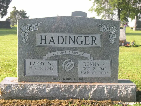 A grey granite upright monument with a raised name panel, floral corners, an epitaph banner and a marriage emblem between two name tablets
