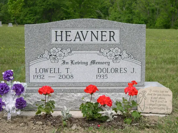 A grey granite upright monument with a raised name panel and a carved floral swag, with a small angel keepsake stone set among the plantings