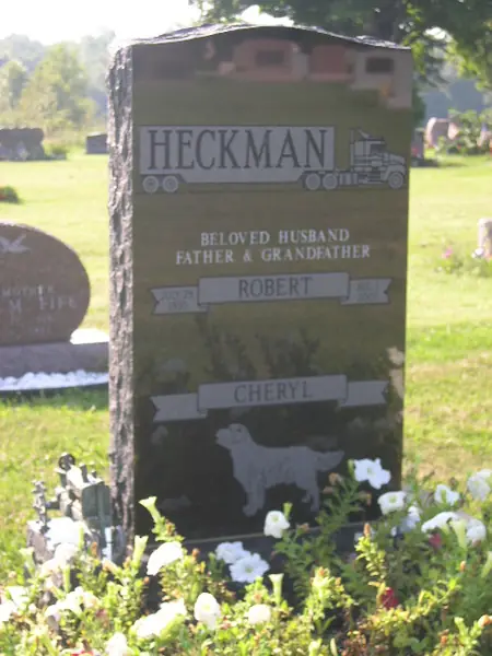 A dark granite upright monument with rough-hewn side edges, an etched semi-truck at the top, a relationship line, two name banners and a carved dog below