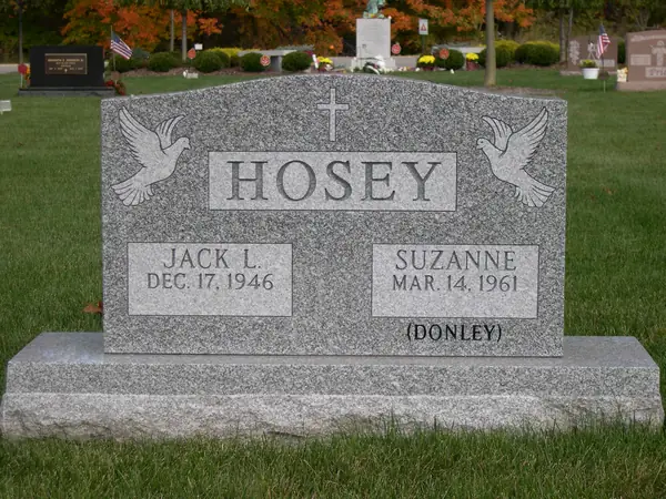 A light grey granite upright monument with doves carved at both shoulders, a cross above the raised name panel and a maiden name cut below