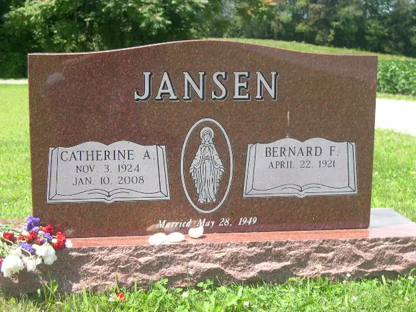 A red granite upright monument with a shaped top, open books flanking a Madonna relief and a marriage date cut below, on a rock-pitched base