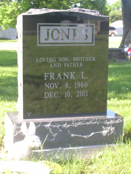 A polished black granite upright monument with a rough-hewn top edge, a raised name panel and a relationship line above the name
