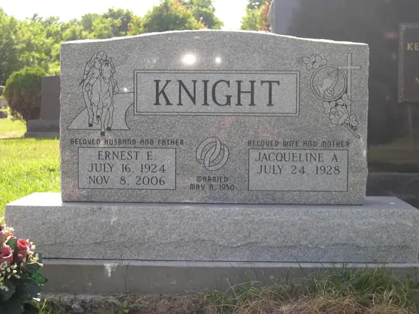 A grey granite upright monument with praying hands carved at one shoulder and a cross with roses at the other, and relationship lines above each name tablet