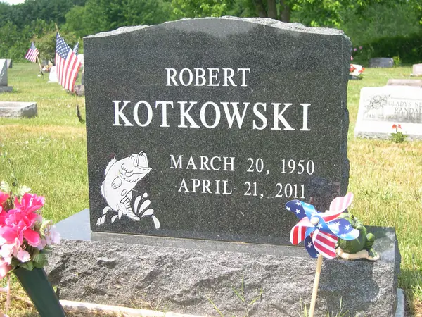 A polished dark grey granite upright monument with a rough-hewn top edge, the full name and dates in raised lettering and a leaping bass etched below
