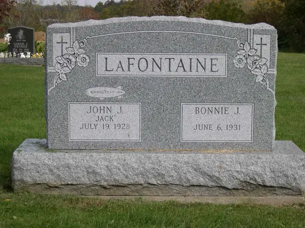 A light grey granite upright monument with a rough-hewn top, carved crosses and flowers at both shoulders and a raised name panel above two name tablets