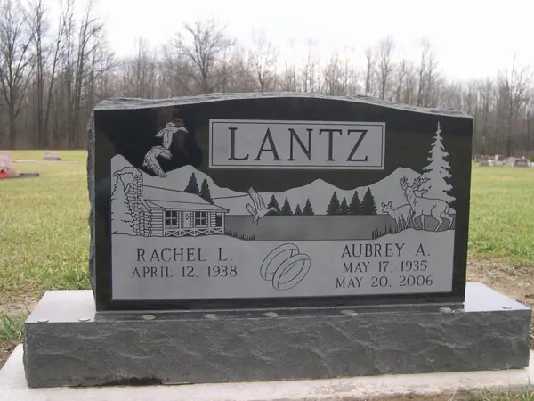 A polished black granite upright monument with an etched cabin, mountains and deer scene, a raised name panel and a wedding-ring emblem between the name tablets