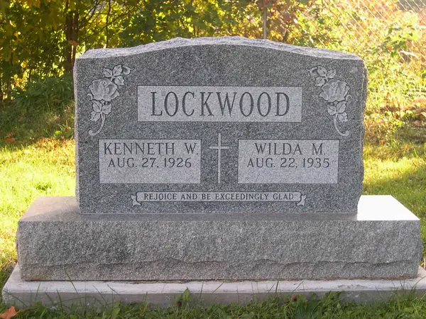 A grey granite upright monument with a raised name panel, roses carved at both upper corners, a cross between the name tablets and an epitaph banner below