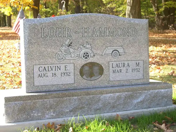 A grey granite upright monument carrying an etched touring motorcycle and trailer, with a small oval portrait between the two name tablets