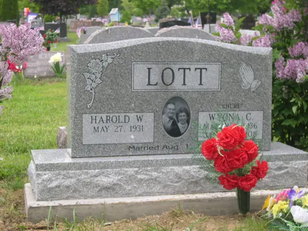 A light grey granite upright monument with a raised name panel, a rose vine at one shoulder and praying hands at the other, and an oval portrait between the name tablets