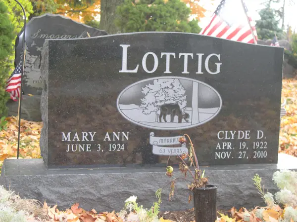 A dark granite upright monument with a raised name panel and an oval etched farm scene of a barn and grazing cattle, with a marriage plaque between the names