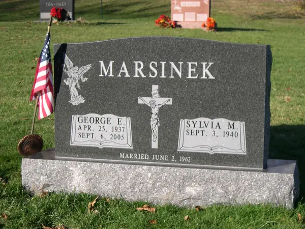A grey granite upright monument with an angel carved at one shoulder, a crucifix on an open book between the name tablets and a marriage date across the foot