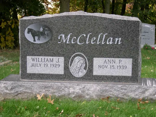 A grey granite upright monument with the family name in script, an oval etched horse and sulky above it and a Madonna relief between the two name tablets