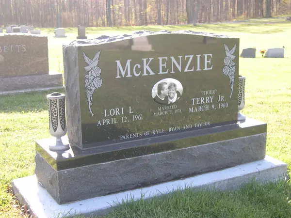 A polished dark granite upright monument with carved fuchsias at both shoulders, an oval etched portrait between the name tablets and matching granite vases