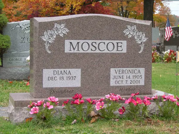 A rose granite upright monument with carved lilies at both upper corners, a raised name panel and relationship lines beneath each of the two name tablets