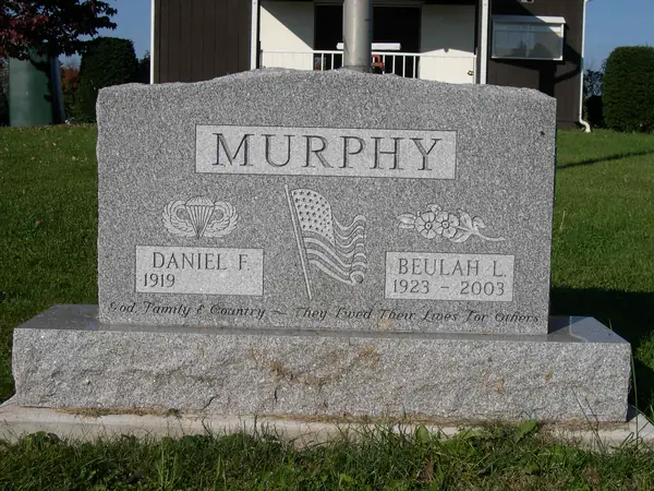 A grey granite upright monument with a raised name panel, carved paratrooper wings, a furled American flag and flowers, and an epitaph across the foot