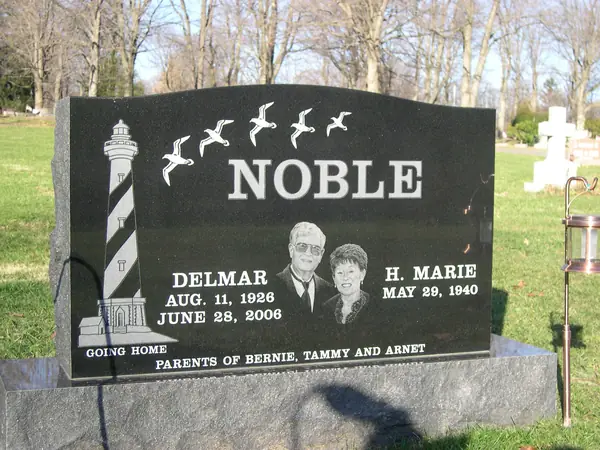 A polished black granite upright monument with an etched lighthouse and gulls in flight, two etched portraits between the name tablets and an epitaph across the foot