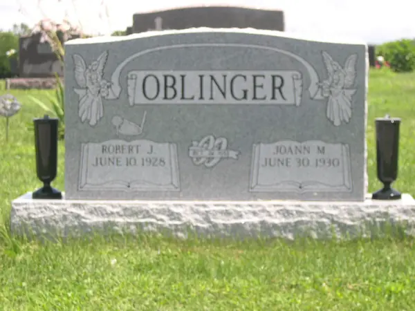 A light grey granite upright monument with angels carved at both shoulders, a raised name panel on a scroll and an open book between the name tablets