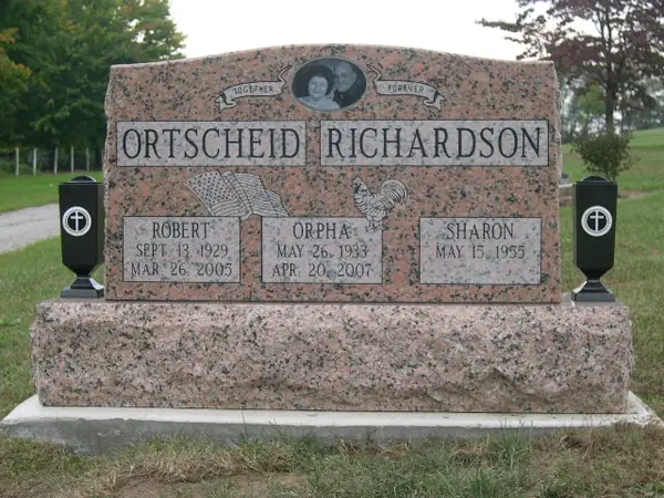 A rose granite upright monument carrying two family names, an oval double portrait on a banner above, three name tablets and a carved rooster, with granite vases either side