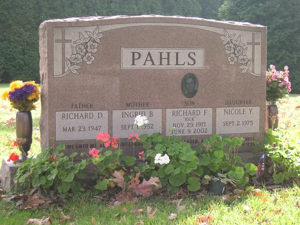A rose granite upright monument with a rough-hewn top, carved crosses and roses at both shoulders, an oval portrait and four name tablets labelled by relationship
