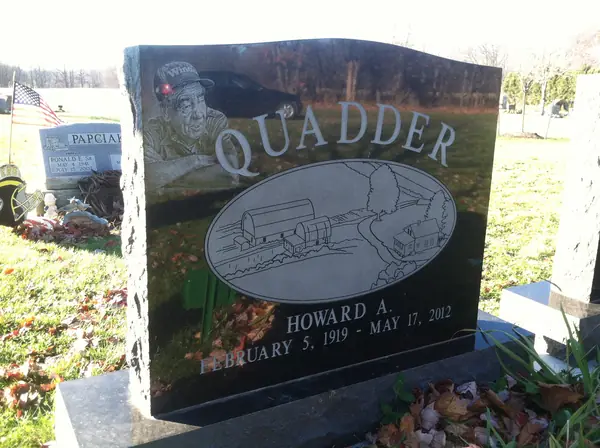 A polished black granite upright monument with rough-hewn side edges, an etched miner's portrait and an oval mining scene above the name and dates