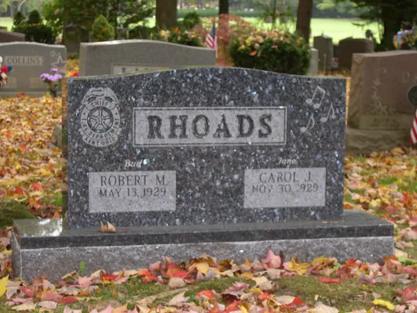 A dark speckled granite upright monument with a carved police badge at one shoulder and musical notes at the other, and nicknames above each name tablet