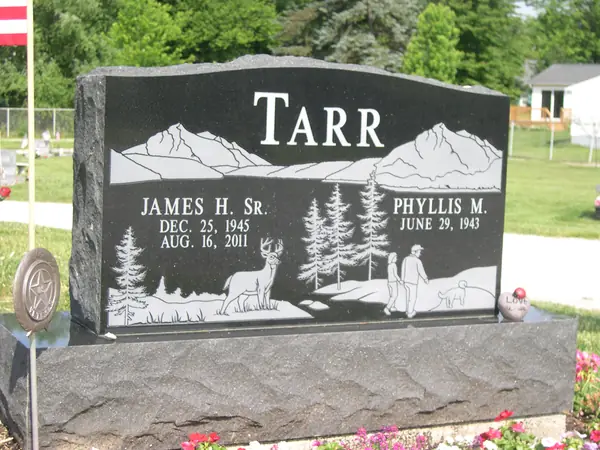 A polished black granite upright monument with a wide etched scene of mountains, pines, a deer and a walker with a dog, on a rock-pitched base