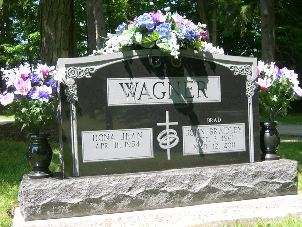 A polished dark granite upright monument with carved scrollwork at the upper corners, a raised name panel and a cross between the name tablets, with bronze vases either side