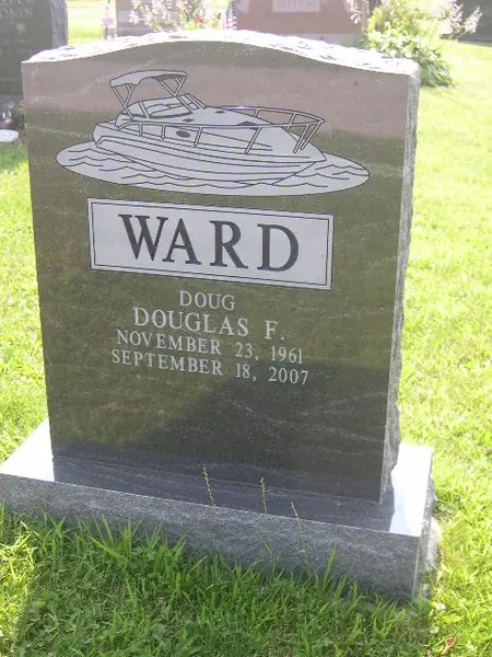 A dark granite upright monument with an etched cabin cruiser above a raised name panel, and a nickname above the full name