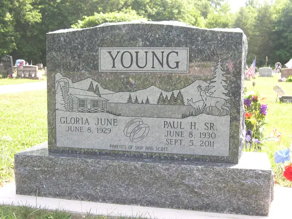 A grey granite upright monument with a rough-hewn top, an etched cabin, pines and deer scene, a raised name panel and a wedding-ring emblem between the names