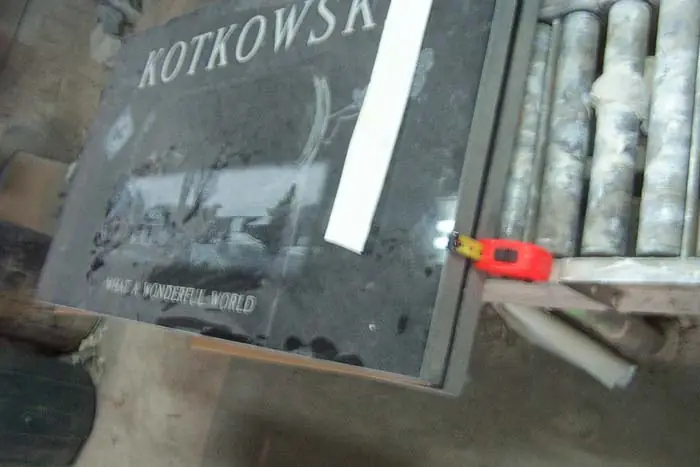 A dark granite monument with an etched scene and a surname standing in the workshop, a straightedge and tape measure resting against it and rolls of stencil material behind.