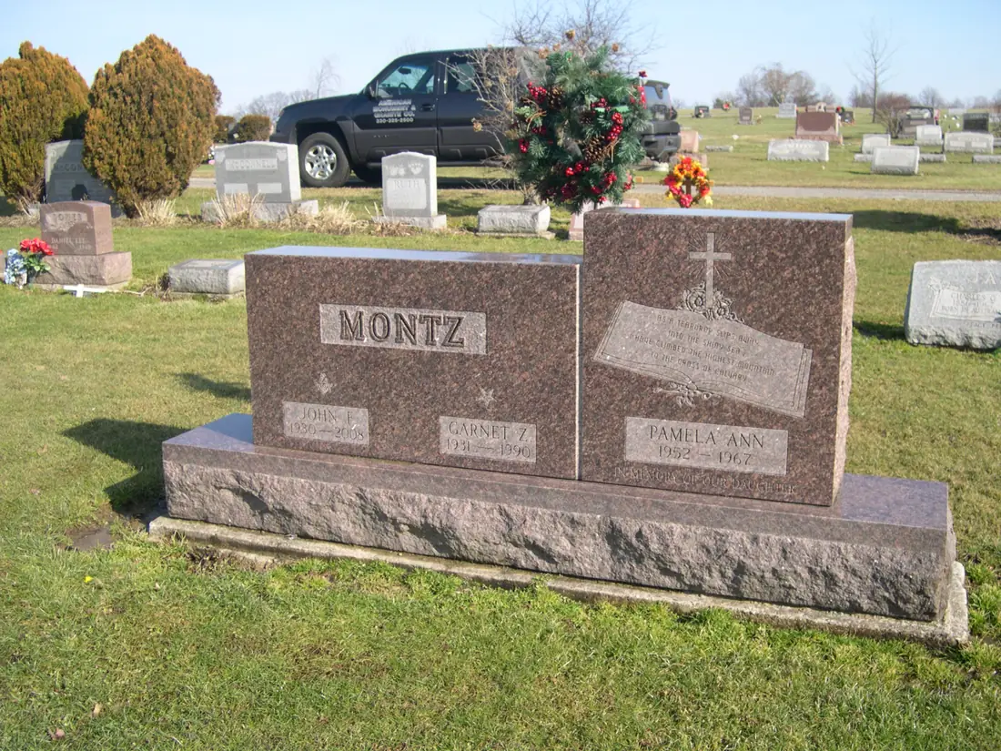 A rose granite monument with a carved cross panel set on a rough granite base in a cemetery, with a pickup truck and other memorials behind.