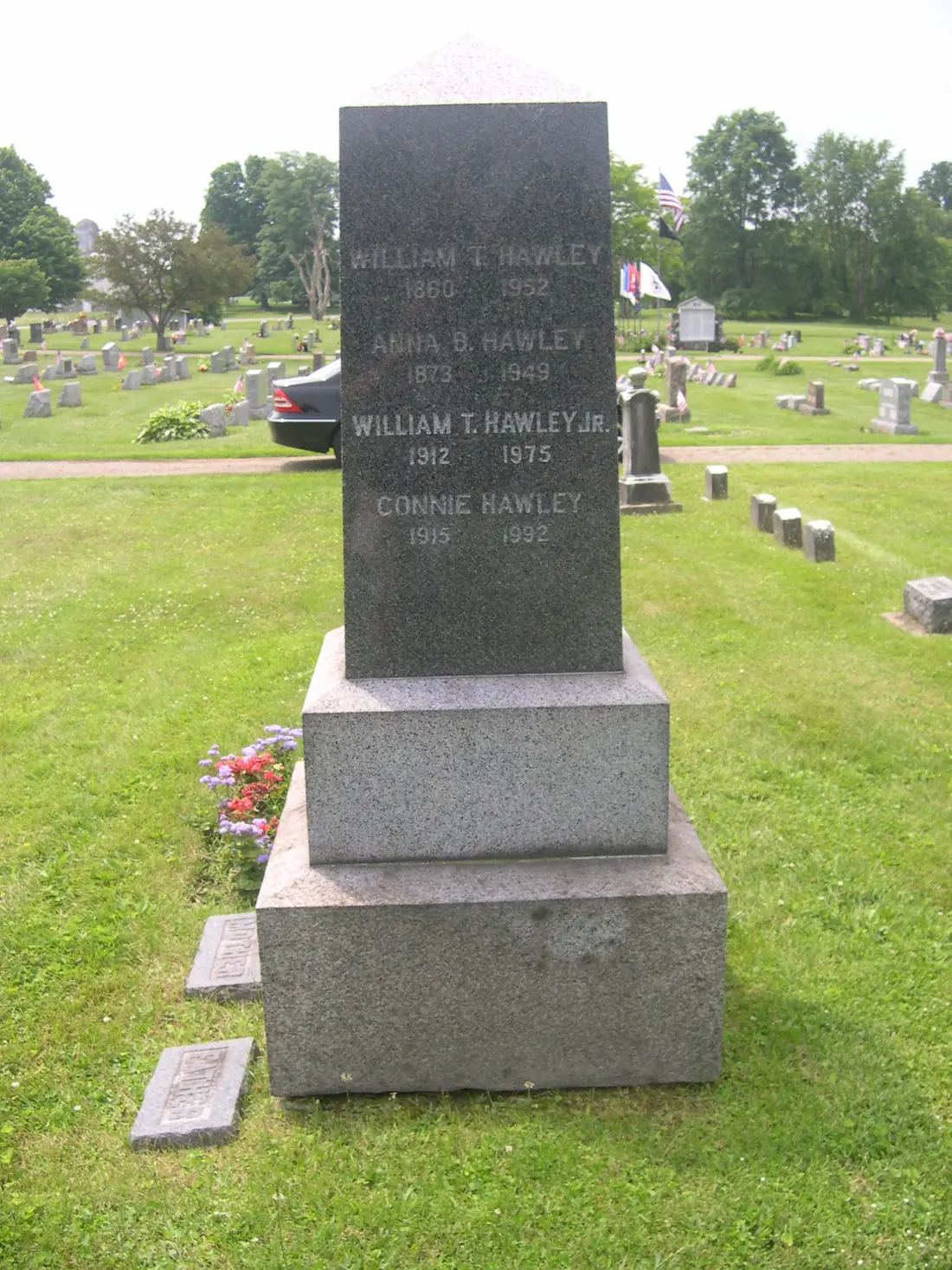 A tall dark granite obelisk on a pink granite base in a cemetery, one face inscribed with four family names and their dates.