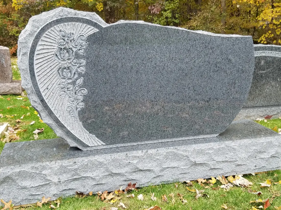A gray granite monument with a carved heart-shaped panel of roses and a fan of rays at one end, standing on a rough-hewn base with autumn trees behind.