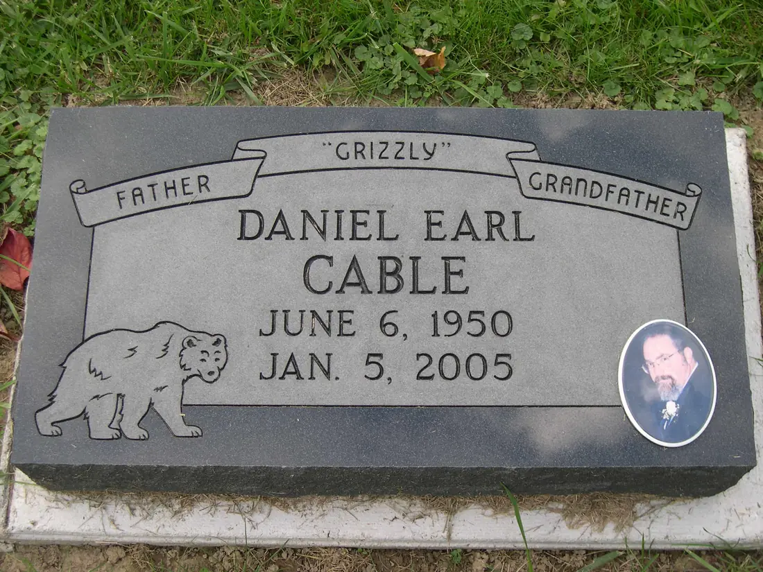 A flat granite marker with the words Father and Grandfather on banners either side of a nickname in quotation marks, above a name and two dates, with a bear etched at one corner and a porcelain photograph at the other.