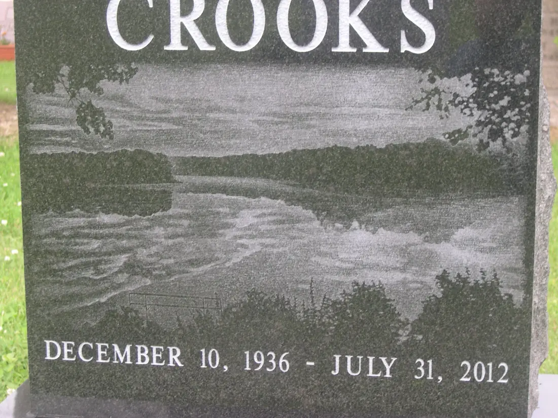 A black granite monument with a lake and treeline laser-etched across it in fine gray detail, a surname above and two dates below.