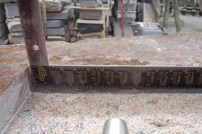 A steel square laid along the edge of a red granite slab on a workbench, with stone and equipment stacked behind it.