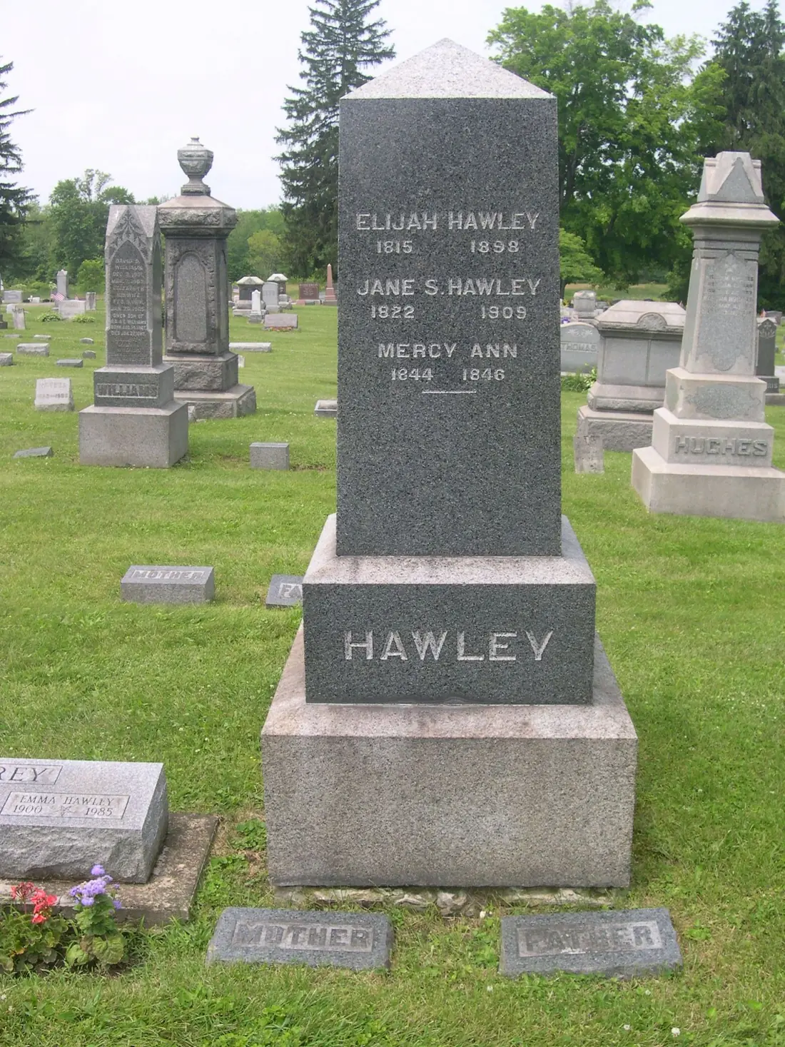 A tall dark granite obelisk on a pink granite base in a cemetery, three names and their nineteenth-century dates still crisply legible, with weathered pale marble monuments standing behind it.