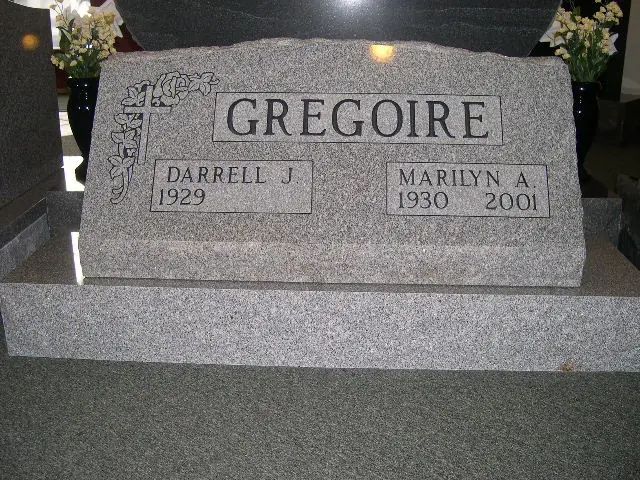 A finished gray granite monument standing indoors in the shop, inscribed with a surname and two given names, one with a birth date only and one with both dates.