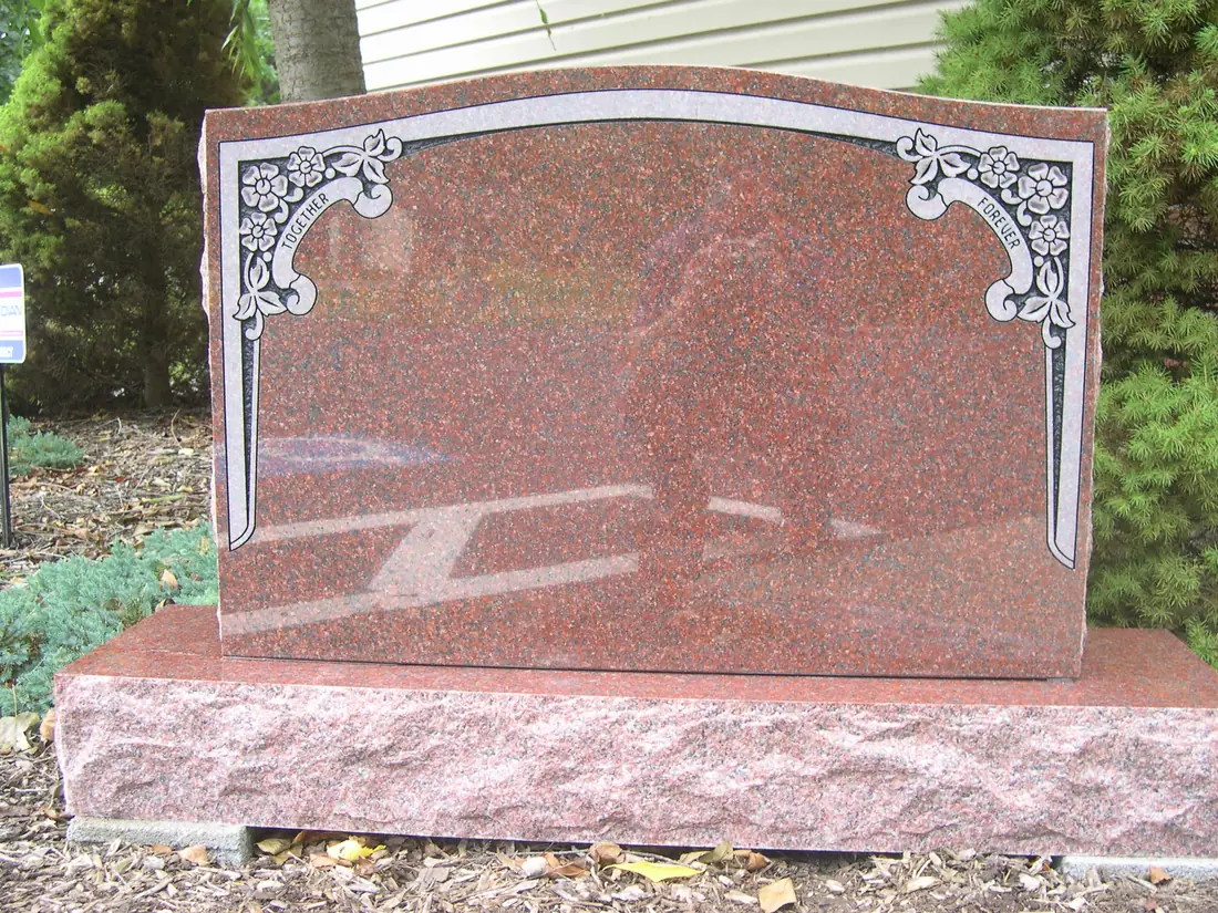 A red granite upright monument with an arched top and ornate floral corners carved in a lighter frosted finish, standing on a rough-cut base.