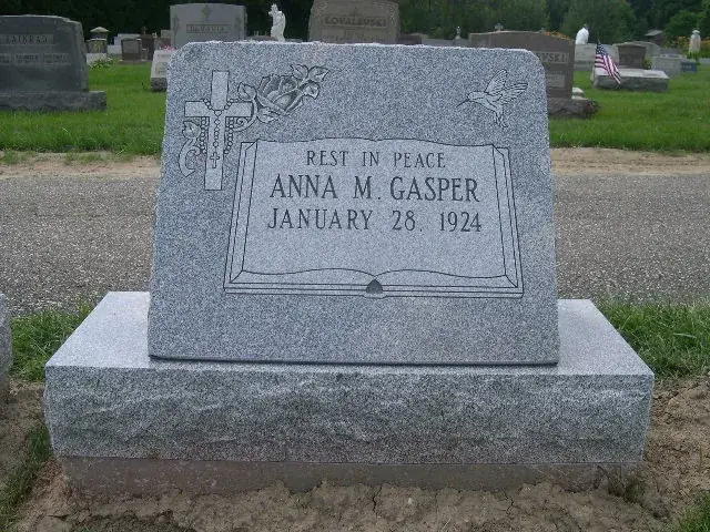 A gray granite slant marker carved as an open book, reading Rest In Peace above a single name and one date, with a cross and floral carving at the corners.